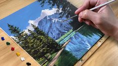 a person is holding a paintbrush and painting a landscape on a piece of wood