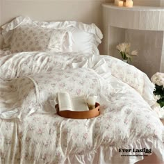 an open book and some flowers on a bed with white comforter, pillows and sheets