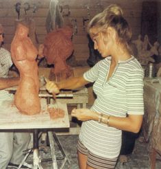 two people working on clay sculptures in a studio with one woman standing and the other man sitting