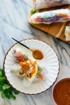 two plates with rolls and dipping sauces on them next to a cutting board full of vegetables