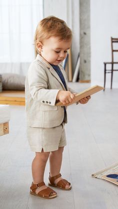 a little boy in a suit and sandals standing on the floor holding a book while looking at it