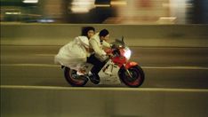 a man and woman riding on the back of a red motorcycle down a city street