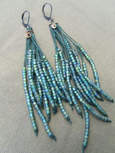 blue and green seed beads are hanging from hooks on a gray fabric background with copper accents