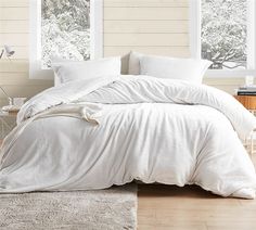 an unmade bed with white sheets and pillows in front of two large windows on a wooden floor