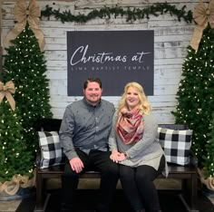 a man and woman sitting on a bench in front of a christmas sign with trees