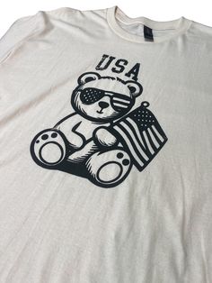 Get into the patriotic spirit with our USA Teddy Bear Design Cotton T-Shirt. Made with a Gildan 100% Heavy Cotton T-shirt, this shirt is both comfortable and durable, perfect for everyday wear. The design features an adorable teddy bear holding an American flag, screen printed onto the front of the shirt. Show off your love for the USA with this fun and festive tee, perfect for Fourth of July celebrations, or simply to show your American pride all year round. Available in a range of sizes, this shirt is a must-have for anyone who loves the U.S.A. Made In Usa Crew Neck T-shirt For Veterans Day, Patriotic 4th Of July Streetwear T-shirt, American Crew Neck Pre-shrunk T-shirt, Veterans Day Flag Print Crew Neck T-shirt, Memorial Day Graphic Tee Made In Usa, White Patriotic T-shirt With Screen Print, Patriotic T-shirt For Independence Day Streetwear, Patriotic T-shirt For 4th Of July Streetwear, Patriotic T-shirt Made In Usa For Veterans Day