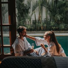 a man and woman sitting next to each other in front of a pool drinking wine