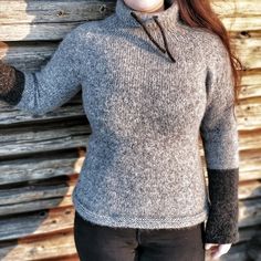 a woman with red hair wearing a gray sweater and black pants standing in front of a wooden wall