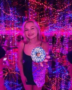 a woman is holding a doughnut in front of some colorful lights at night time