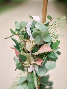 an arrangement of greenery is displayed in a vase on a table with a wooden stick