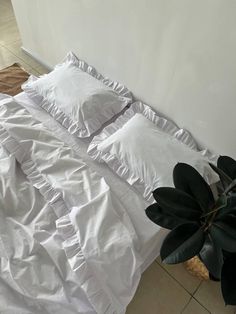 an unmade bed with white sheets and pillows next to a potted plant on the floor