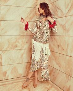 a woman standing in front of a wall wearing a white and gold dress with red flowers