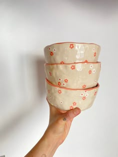 a hand holding three bowls in front of a white wall with red flowers on them