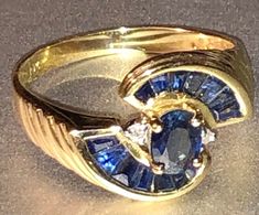 This lovely art deco revival 1970s-80s ring, is stamped 10K yellow gold, and boasts a lovely retro design with a center sapphire surrounded by channel set stones and a couple of little satellite diamonds. I believe the sapphires to be natural and untreated. The ring is about a size 7 and would be great as a birthstone ring or alternative engagement ring. Weighing approximately 2.7g total, it's in very good vintage condition with no damage or repair. Make sure to view all photos and zoom in for d Gold Oval Sapphire Ring In Art Deco Style, Vintage Multi-stone Oval Sapphire Ring, Vintage Oval Multi-stone Sapphire Ring, Vintage Multi-stone Sapphire Ring In 14k Gold, Vintage Gold Sapphire Ring With Accent Stones, Vintage Gold Sapphire Ring With Emerald Cut, Vintage Sapphire Rings Stamped 14k, Art Deco Sapphire Ring, Art Deco Revival