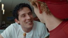 a man is smiling and talking to another man in a red baseball cap while he eats an ice cream cone