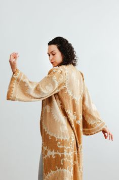 This semi-fitted duster was made from an antique hand-embroidered woven cotton Suzani, a textile traditionally used as wall hangings or in the yurts of nomadic people in Central Asia. An original aloka design, we prepared the vintage material with our signature multi-step shampoo and softening process. In our Athens, Georgia-based studio, our designers created this A-line semi fitted jacket with a plunging neckline and puffed bell sleeves. Served bound seams on the interior showcase the hand-stitching. Thick trim along the bottom of the jacket and sleeves give it a luxurious look and drape. Each textile bares memories and marks of the hand and past. Each piece is a one-of-a-kind garment. We carefully curate and prepare these beautifully preserved textiles and embrace imperfections, mending Traditional Beige Kaftan For Festivals, Traditional Brown Kimono For Festival, Traditional Cotton Ikat Print Kimono, Traditional Cotton Kimono With Ikat Print, Beige Embroidered Kimono With Kimono Sleeves, Traditional Brown Kaftan With Kimono Sleeves, Traditional Long Brown Kimono, Traditional Beige Cotton Kimono, Traditional Long Sleeve Kimono With Natural Dye