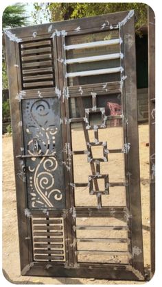 two metal doors with designs on them in the sand and trees behind them, one door is open