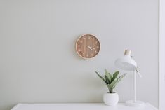 a white desk with a clock on the wall next to a potted plant and lamp