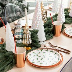 the table is set for christmas dinner with gold and white plates, silverware, green napkins and trees