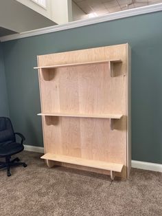 a wooden book shelf sitting in an empty room next to a black chair and gray wall