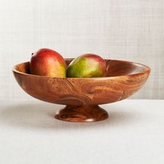 two apples in a wooden bowl on a white tableclothed surface with a vase behind it