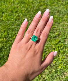 a woman's hand holding an emerald ring on top of her finger in the grass