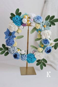 a crocheted wreath with blue and white flowers sits on a stand next to a window