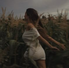 a woman in a white dress walking through a corn field with her hair blowing in the wind