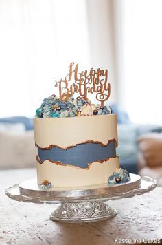 a birthday cake with blue and white frosting is on a glass platter in front of a window