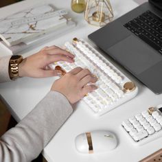 a person typing on a white keyboard with gold accents at a desk in front of a laptop