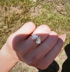 a person's hand holding an engagement ring with two diamonds on it and grass in the background