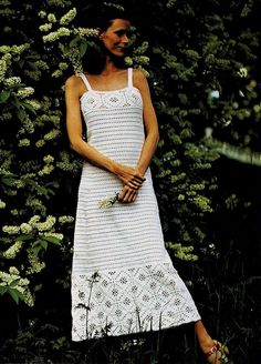 a woman in a white dress standing next to some bushes and flowers with her hands on her hips