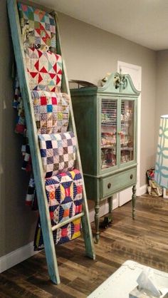 a room with a ladder and quilts on the wall next to a dresser in it