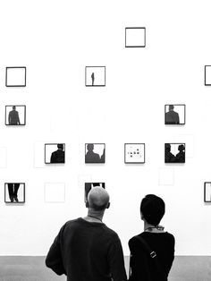 two people standing in front of a white wall with many square photos on it