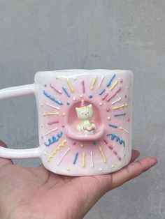 a hand holding a white coffee mug with colorful sprinkles and a cat in it