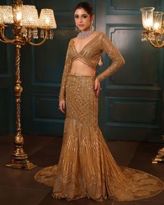 Metallic gold fishcut lehenga with all-over sequin, crystal, and cutdana embroidery with a flowing trail at the back. Paired with a delicate full sleeves blouse with hanging tassel detailing and embroidered dupatta in net.DELIVERY TIMEPlease allow 8-12 weeks for your outfit to arrive.FABRIC DETAILSNetProfessional cleaning only. Gold Pre-draped Saree With Resham Embroidery For Evening, Glamorous Floor-length Sharara With Resham Embroidery, Glamorous Semi-stitched Lehenga With Sheer Dupatta, Glamorous Evening Sharara With Dupatta, Glamorous Gown With Sequins In Saree Style, Glamorous Hand Embellished Diwali Dresses, Glamorous Embellished Semi-stitched Lehenga, Glamorous Hand Embellished Semi-stitched Lehenga, Glamorous Gold Long Sleeve Gown