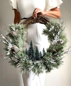 a woman is holding a wreath with pine trees and evergreens on the front, while wearing a white dress