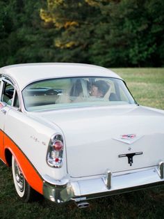 an orange and white vintage car is parked in the grass with two people sitting in it