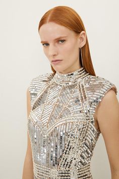 a woman with long red hair wearing a silver dress and earrings, standing in front of a white wall