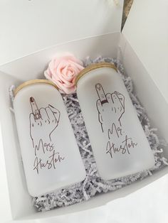 two white jars with writing on them are in a box next to a pink rose