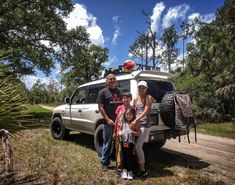 a family poses in front of their vehicle on a dirt road with trees and grass