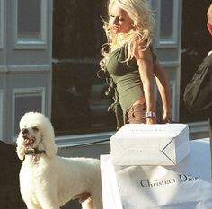 a white poodle standing next to a woman in a green shirt and brown pants