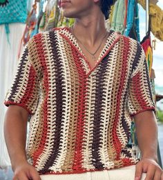 a man wearing a crochet shirt standing in front of some clothes hanging up