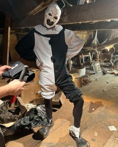 a person in a clown mask and black pants standing on a wooden floor with other people around