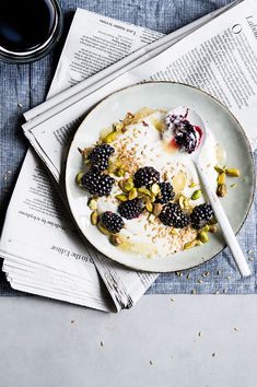 a white plate topped with fruit and granola next to a stack of newspapers on top of a table