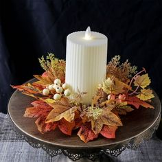 a white candle sitting on top of a cake plate filled with autumn leaves and berries