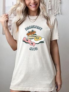 Introducing our Breakfast Club Tee, a must-have addition to your casual wardrobe! Embrace your love for all things breakfast with this charming retro graphic t-shirt. The front design features delightful cartoons of pancakes, eggs, and bacon, creating a nostalgic and whimsical vibe. Crafted with care, this shirt is perfect for brunch enthusiasts, pancake aficionados, and anyone who appreciates a touch of vintage charm. Made from high-quality materials, this Retro Brunch Shirt exudes a hippie-inspired retro aesthetic, blending style and comfort effortlessly. Stand out from the crowd and celebrate the most important meal of the day in true fashion with our Breakfast Shirt - it's a brunch lover's dream come true! Overview -These t-shirts have-ribbed knit collars to bolster shaping. -The shoul Cotton T-shirt With Letter Print For Brunch, White Crew Neck T-shirt For Brunch, Relaxed Fit Cotton Shirt For Brunch, Cotton Shirt With Relaxed Fit For Brunch, Funny Print Graphic Tee For Brunch, Funny Print Short Sleeve T-shirt For Brunch, Casual Screen Print T-shirt For Brunch, Short Sleeve Tops With Funny Print For Brunch, Short Sleeve Screen Print Tops For Brunch