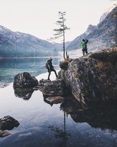 two people are standing on the edge of a mountain overlooking a body of water with mountains in the background