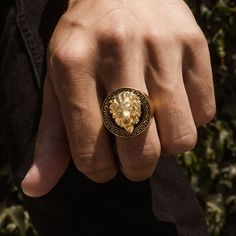 a man wearing a ring with a lion head on it's middle finger, in front of some bushes