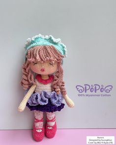 a crocheted doll with pink hair and purple dress is posed against a white wall